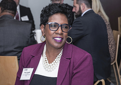 Ann Levett, a Black woman with short black hair, wearing a purple blazer and white and black top with pearl necklace and gold earrings