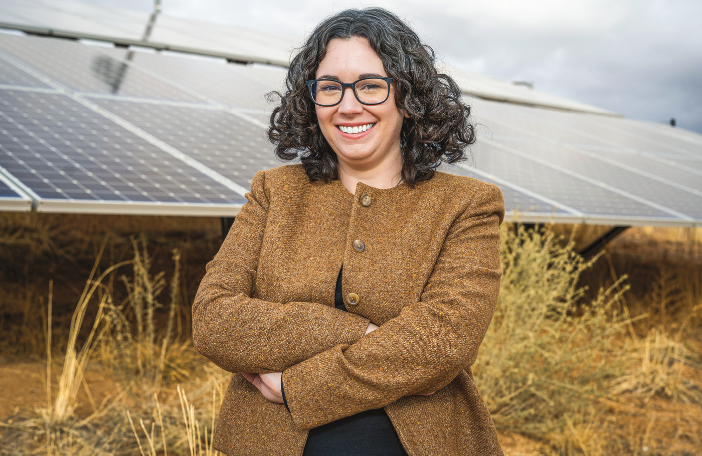 A woman with curly brown-gray shoulder length hair in front of a solar panel on a field wearing a brown jacket