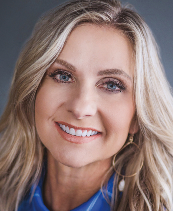 A white blonde woman with blue eyes smiling wearing a blue collared shirt