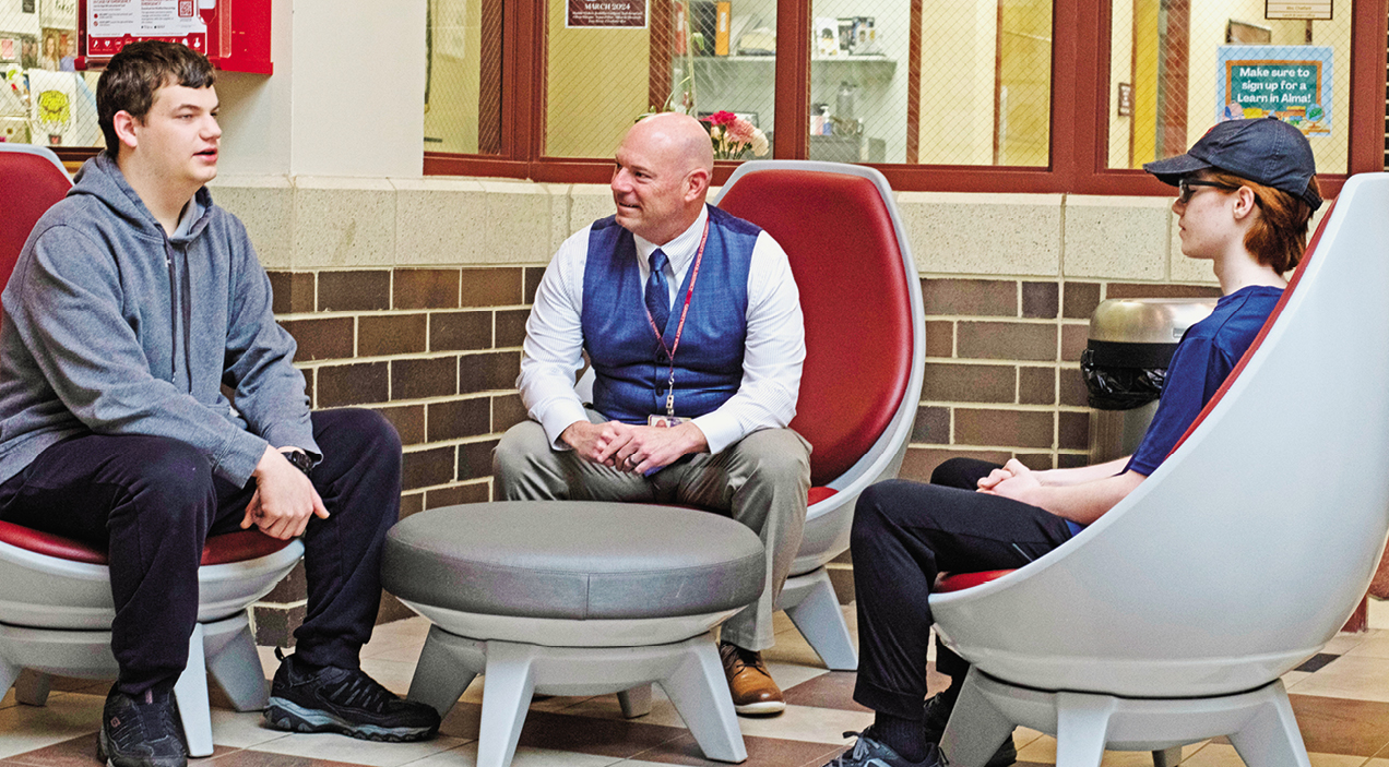 An adult talks to two students; they are all seated