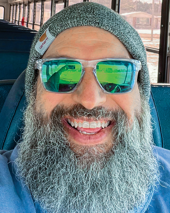 A man wearing a gray beanie, with a long, thick gray beared smiling and wearing sunglasses