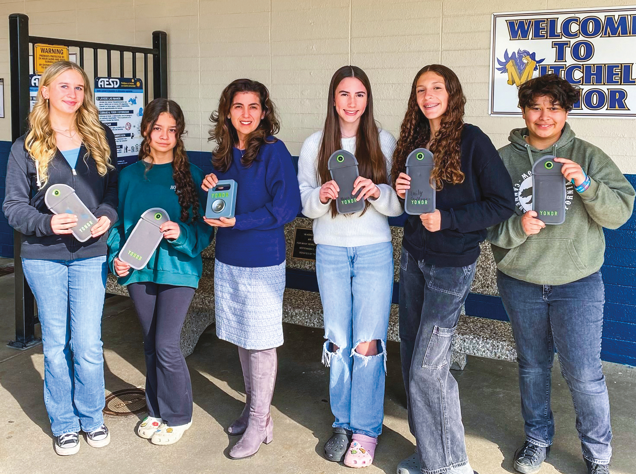 A group of teenagers with an adult in the middle all holding pouches that lock away their cell phones