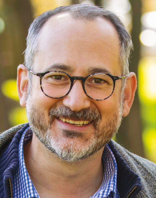 A White Man wearing round glasses and a gray sweater, with gray hair and beard