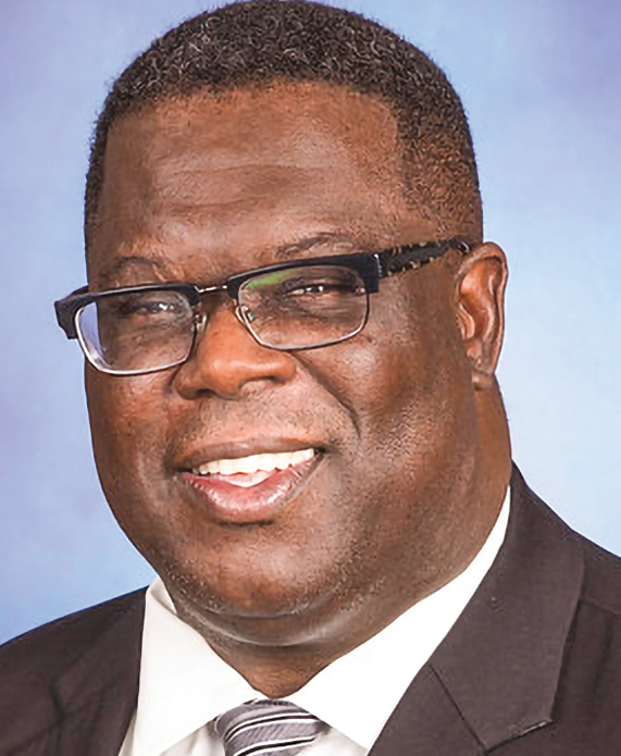 A Black man with short black hair wearing black-rimmed glasses and a black suit with white shirt