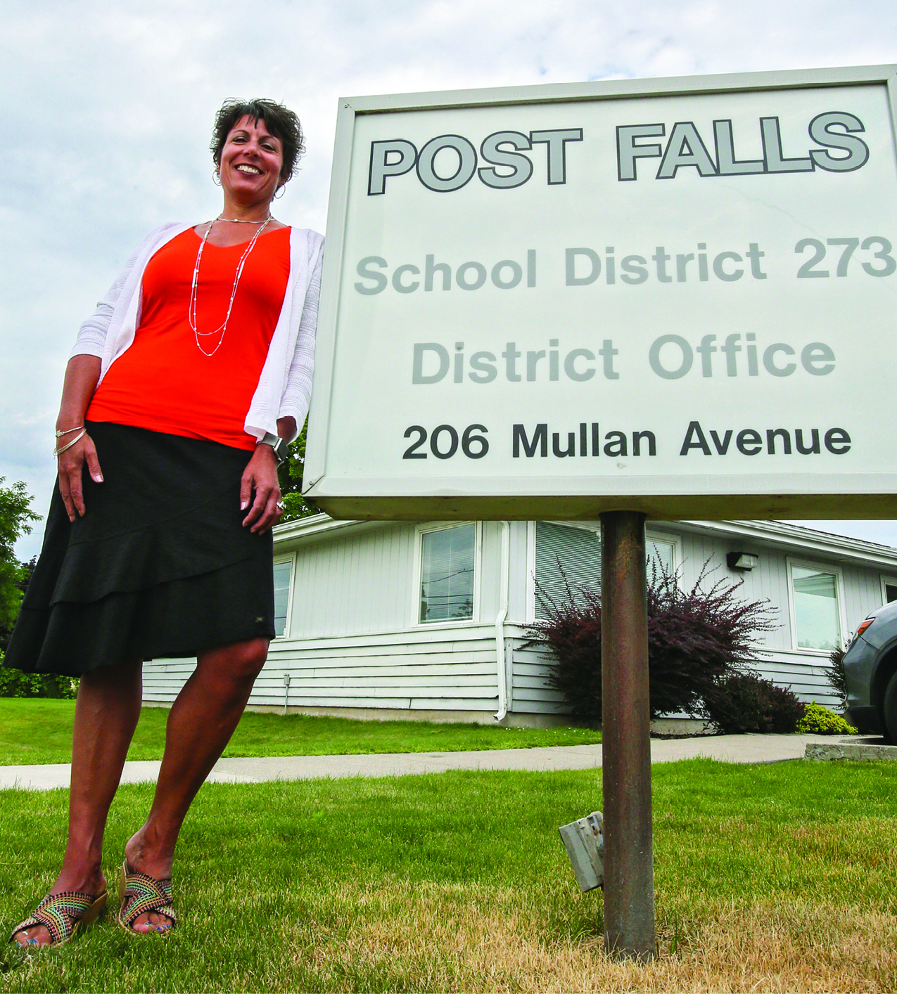 Dena Naccarato, a woman in red blouse and black skirt with white cardigan, leaning against a sign that says Post Falls School District 273 District Office 206 Mullan Avenue