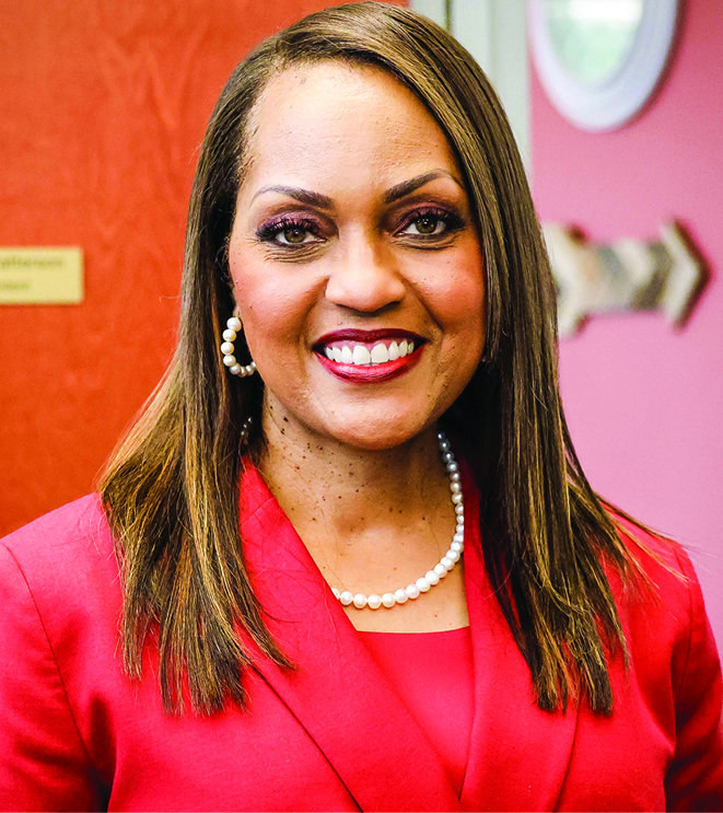 Paula Patterson, a Black woman with straight long brown hair smiling wearing pearls and a red suit